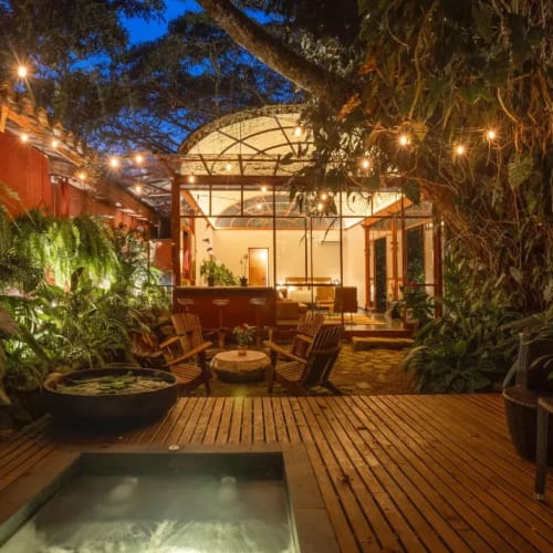 A cozy outdoor hotel area at night with warm lighting, lush green plants, wooden furniture, and a glass-walled building in the background.
