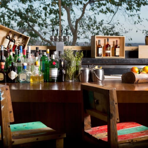 Ein Barbereich mit verschiedenen Alkoholflaschen, Gläsern und frischen Früchten auf einer Holztheke, mit Stühlen und Blick auf Bäume draußen.