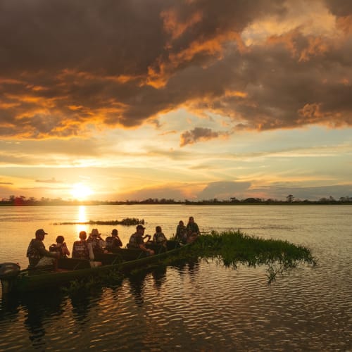 Eine Gruppe von Menschen sitzt auf einem Boot auf einem Fluss bei Sonnenuntergang, vor einem dramatischen Himmel mit dunklen Wolken.