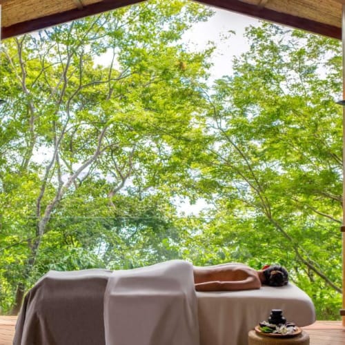 Eine Person erhält eine Spa-Behandlung im Freien auf einer Massageliege mit Blick auf grüne Bäume.