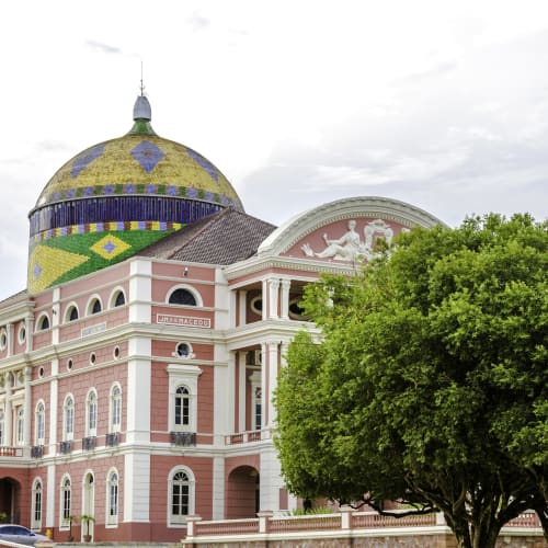 A colorful opera house with a patterned dome, pink and white facade, and a lush green tree in front, set against a cloudy sky in Manaus, Amazonas.