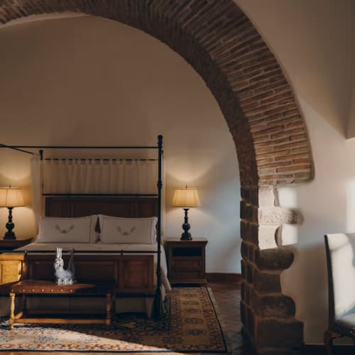 Ein Schlafzimmer im Kolonialstil im Palacio Nazarenas, einem Belmond Hotel in Cusco, mit einem hölzernen Himmelbett mit bestickten Kissen, Nachttischen mit Lampen, einem verzierten Teppich und warmem Licht, das einen Steinbogen hervorhebt.