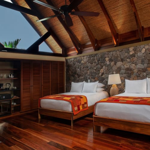 Zweibetten mit weißen Bettlaken und orange-roten Decken in einem gemütlichen Raum mit Steinwand, Holzdecke und einer Lampe auf einem Nachtschränkchen.