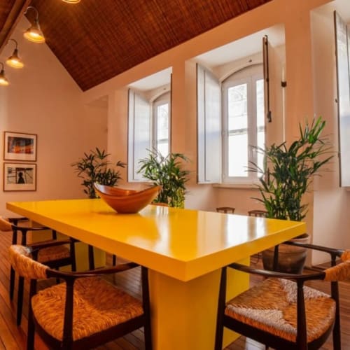 Cozy dining area in Pousada Literária de Paraty featuring a bright yellow table surrounded by woven chairs, wooden floors, and large windows letting in natural light.