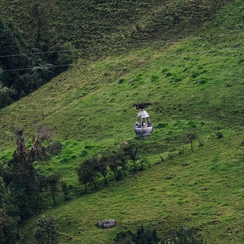 Eine Seilbahn-Gondel schwebt über einer grünen bergigen Landschaft mit verstreuten Bäumen und Felsen.