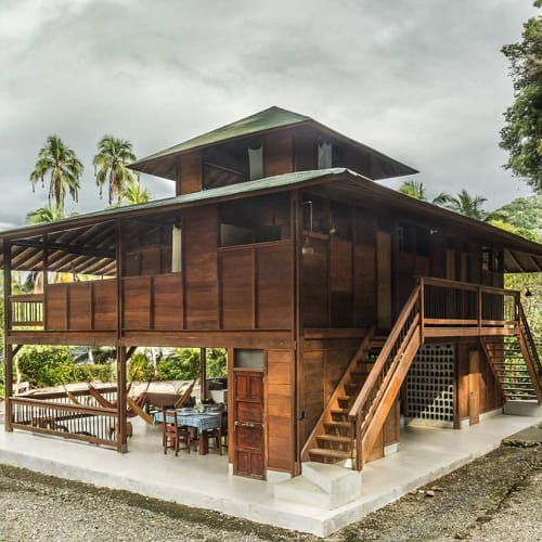 Ein traditionelles erhöhtes Holzhaus mit zwei Treppen, die zur oberen Ebene führen, umgeben von tropischen Palmen und Grün unter einem bewölkten Himmel.