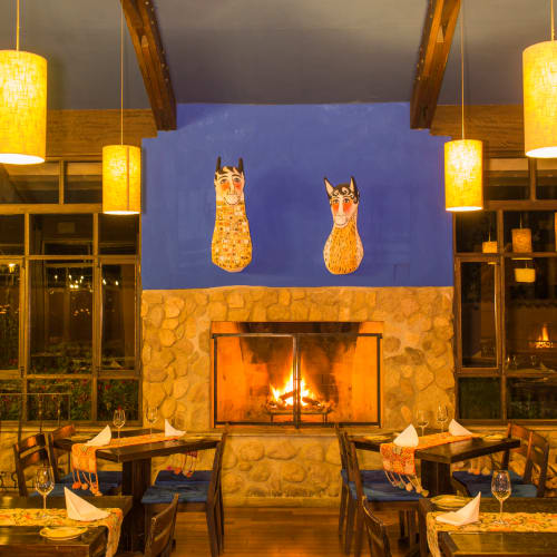 Gemütliches Restaurant mit Holzstühlen und gedeckten Tischen vor einem steinernen Kamin mit Feuer, umgeben von großen Fenstern und warmen Deckenleuchten.