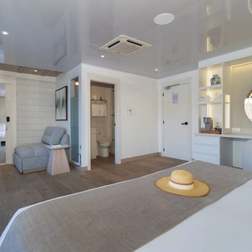 Geräumiges modernes Hotelzimmer mit einem großen Bett, grauem Sessel mit Beistelltisch, durch eine offene Tür sichtbarem Badezimmer und einem Schminkbereich mit rundem Spiegel und Beleuchtung.