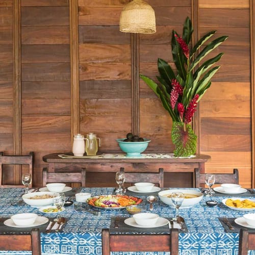 A dining table set with white bowls, plates, and glasses on a blue patterned tablecloth in a wooden room, with a table in the background holding a turquoise bowl and a vase with red flowers and green leaves.