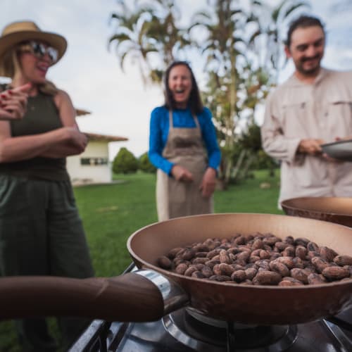 Menschen beim Backen oder Kochen im Freien auf einem Grill, mit Fokus auf einer Pfanne gerösteter Nüsse, im Hintergrund ein Haus und Bäume.