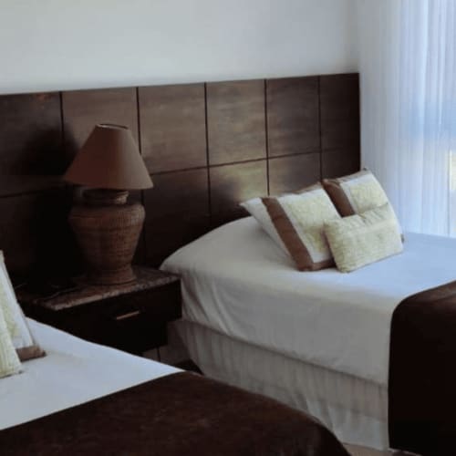 Zwei ordentlich gemachte Betten mit weißen Bettlaken und braunen Decken in einem Hotelzimmer mit großem dunklem Holz-Kopfteil, Nachttischlampe und Meerblick durch ein Fenster mit transparenten Vorhängen.