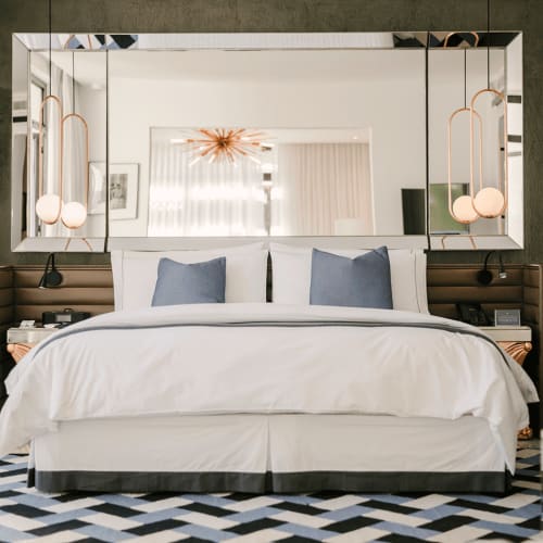 Modernes Schlafzimmer mit einem großen Bett mit weißer Bettwäsche und zwei blauen Kissen, flankiert von braunen gepolsterten Kopfstützen und Hängeleuchten, sowie einem großen rechteckigen Spiegel, der einen Kronleuchter und Fenstervorhänge reflektiert.