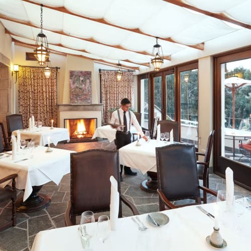 Elegantes Restaurant mit weißen Tischdecken und einem gemütlichen Kamin, Mitarbeiter bereiten den Tisch vor, große Fenster mit Blick auf den Außenbereich mit Sonnenschirmen.