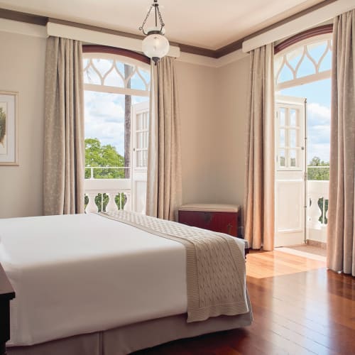 Ein helles Hotelzimmer mit großen Fenstern und Balkontüren, die Tageslicht hineinlassen und auf grün bewachsene Umgebung schauen.