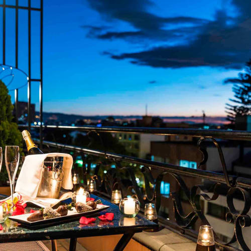 Ein eleganter Außenbereich mit einem Tisch für ein romantisches Abendessen, Kerzen, Rosenblätter und eine Flasche Champagner, mit Blick auf die Stadt bei Sonnenuntergang.