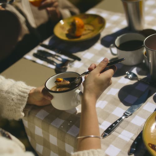Zwei Personen frühstücken im Freien an einem Tisch mit kariertem Tischtuch; eine Person hält eine Tasse mit Brei und einem Löffel, während die andere eine Orangenscheibe isst.