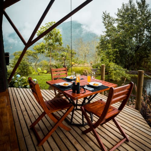 Wooden table set with plates and drinks on a deck overlooking lush green trees and misty mountains in the background.