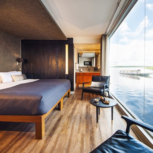 Luxuriöse Suite mit großem Fenster, das auf den Amazonas-Fluss blickt, mit einem Kingsize-Bett, einem kleinen Tisch mit Snacks und Sitzgelegenheit.