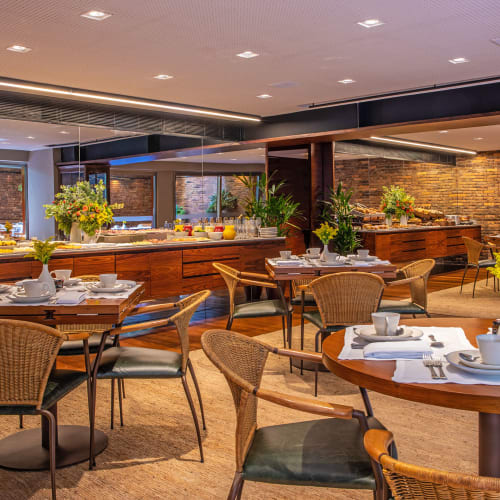 Elegantes Restaurantinterieur mit runden Holztischen, die zum Essen gedeckt sind, Korbsesseln, dekorativen Pflanzen und einem Buffetbereich mit Speisen und Getränken auf einem Holztresen.