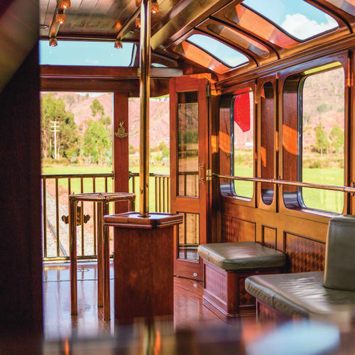 Interior of a wooden train carriage with leather benches and large windows showing green fields and hills outside.