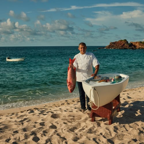 Koch steht an einem sandigen Strand neben einem Holzgestell mit frischem Fisch, hält einen großen roten Fisch, mit Meer und felsiger Küste im Hintergrund.