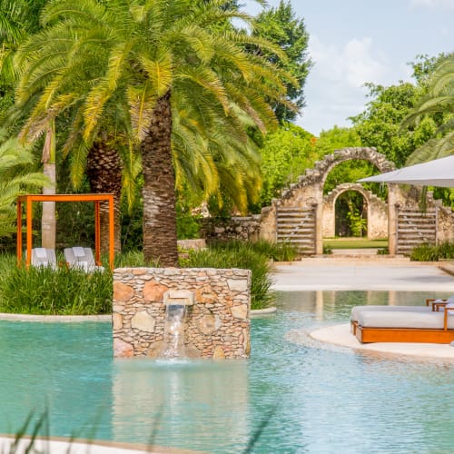 Poolbereich eines Resorts umgeben von üppigen Palmen mit steinigem Wasserfall und Liegen unter großen Sonnenschirmen bei teilweise bewölktem Himmel.