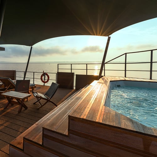 Ein Jacuzzi auf dem Deck eines Bootes oder Kreuzfahrtschiffs bei Sonnenuntergang mit Liegestühlen und Blick auf den Fluss.