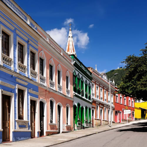 Bunte Kolonialgebäude säumen eine Straße in Candelaria, Bogotá, unter einem strahlend blauen Himmel mit einem Hügel im Hintergrund.