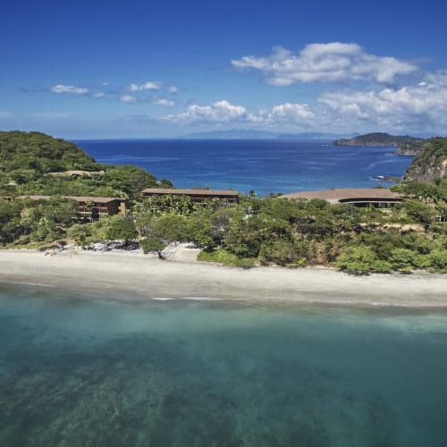 Aerial view of a coastal landscape with a beach, lush greenery, and buildings set among trees under a partly cloudy sky.