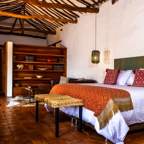 Cozy junior suite with a large bed featuring white linens and red patterned pillows, wooden furniture, terracotta tiled floor, and rustic exposed wooden beams on the ceiling.