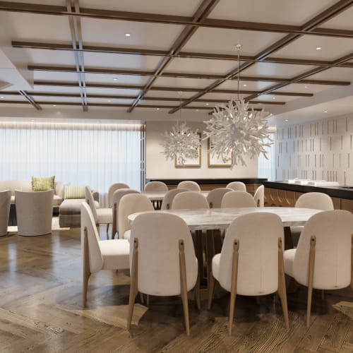 A modern dining room with a large round table surrounded by white upholstered chairs, a green armchair in the foreground, and a seating area with sofas in front of large windows with sheer curtains.