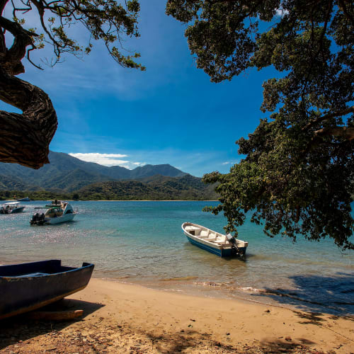 Kleine Boote ankern nahe einem Sandstrand, eingerahmt von großen Baumästen, mit klarem blauem Wasser und entfernten Bergen unter hellem Himmel.