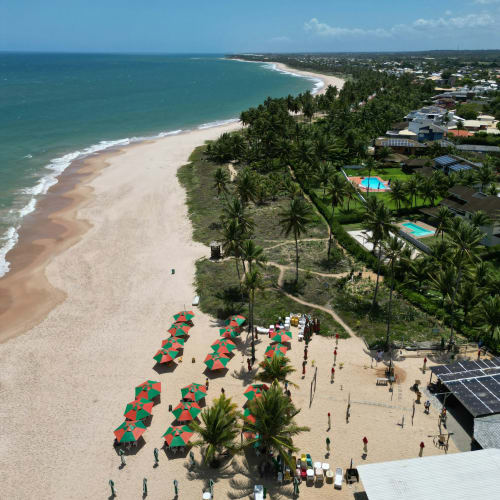 Luftaufnahme des Strandes Praia do Forte in Bahia, Brasilien, mit goldenem Sandstrand, grünen Palmen, bunten Sonnenschirmen und nahegelegener Wohngegend mit Swimmingpools unter klarem blauem Himmel.