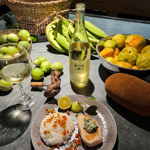 A sunlit table with a plate of rice and grilled fish topped with a green sauce, surrounded by fresh green limes, a bunch of green bananas, a bottle of white wine, a glass of wine, and bowls filled with various fruits including green and yellow citrus.