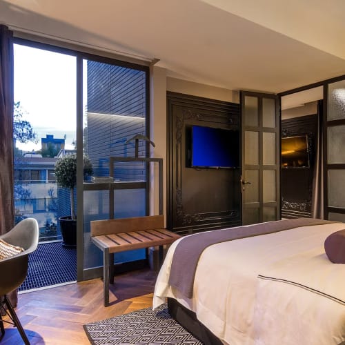 Modernes Hotelzimmer mit großem Bett, dekorativen Kissen, Schreibtisch mit Stühlen und einer Glasschiebetür, die auf einen Balkon mit Blick auf Stadtgebäude öffnet.