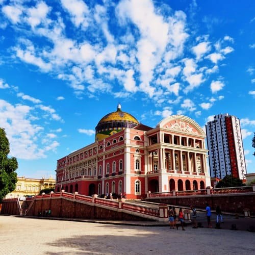 A historic opera house with a pink facade and a golden dome, situated in Manaus, Brazil, under a bright blue sky with scattered clouds.