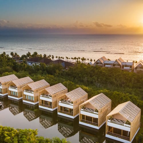 Sonnenuntergang über einer Reihe von modernen hölzernen Strandvillen mit Dachterrassen, die sich in einer Lagune spiegeln, umgeben von dichtem Grün und mit Blick auf das Meer.