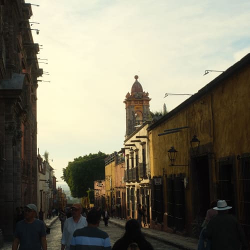 Eine Straßenszene in San Miguel de Allende mit Menschen, die entlang gepflasterter Straßen spazieren, kolonialstillichen Gebäuden und einem Kirchturm im Hintergrund bei frühem Abendlicht.