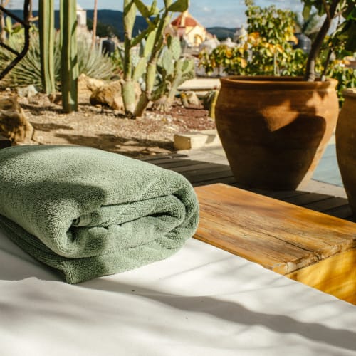 Gefaltetes grünes Handtuch auf einer weißen Spa-Liege im Freien mit Wüstenpflanzen und Tontopf im Hintergrund unter blauem Himmel mit Wolken.