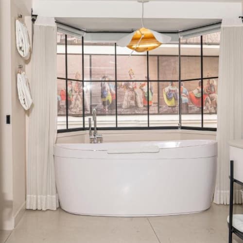 Modernes Badezimmer mit einer weißen freistehenden Badewanne vor großen Erkerfenstern mit einem Wandbild bunter Figuren, dazu weiße Vorhänge und an der Wand befestigte Handtücher.