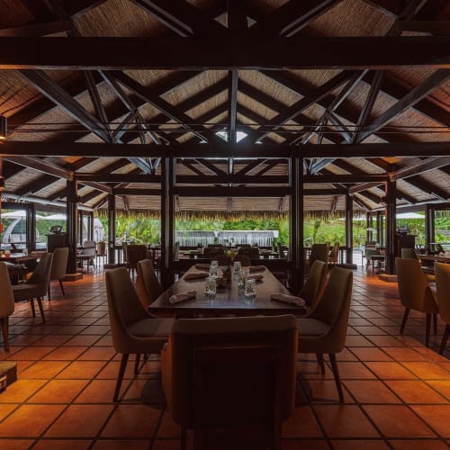 Interior of a rustic restaurant with wooden beams, terracotta tiled floor, and tables set with chairs in a spacious open-air design surrounded by greenery.