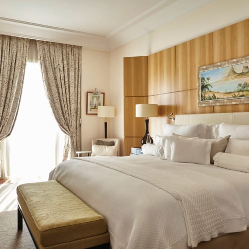 Luxurious hotel room with a large bed, a bench at the foot, and elegant decor, including curtains and paintings, in a high-end setting.