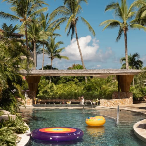 Tropischer Außenpoolbereich mit Palmen, einem Strohdach-Bar, aufblasbaren Schwimmkörpern im Wasser und Liegestühlen mit Sonnenschirmen an einem Luxusresort.
