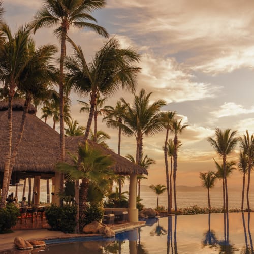 Ein Poolbar mit Strohdach, umgeben von Palmen bei Sonnenuntergang, die sich im ruhigen Infinity-Pool mit Blick auf das Meer im Hintergrund spiegeln.