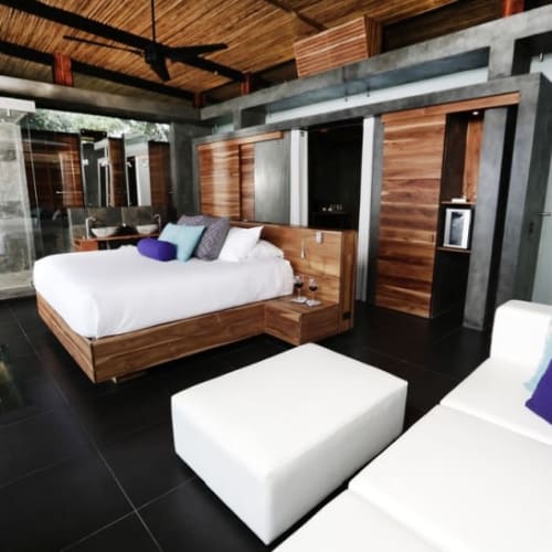 Innenraum eines modernen Hotelzimmers mit einem Holzbett, weißen und lila Kissen, und einem weißen Sofa mit bunten Kissen, im Hintergrund ein Spa-Bereich mit Dusche.