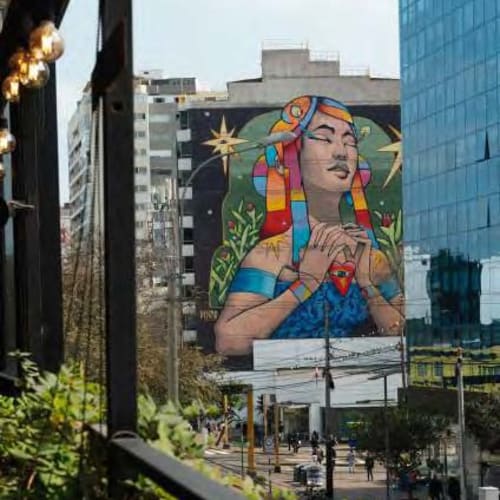 Ein großes Wandbild einer Frau mit bunten geflochtenen Haaren und verschränkten Händen, umgeben von Sternen und Pflanzen, gemalt an einem Stadtgebäude.
