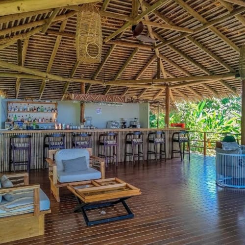 Open-air bar area with a wooden thatched roof, wooden floor, and bar stools lined up at the counter, surrounded by lush green tropical plants.