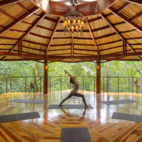 Eine Person praktiziert Yoga in der Krieger-Position in einem offenen Holzpavillon, umgeben von üppigem grünem Wald.