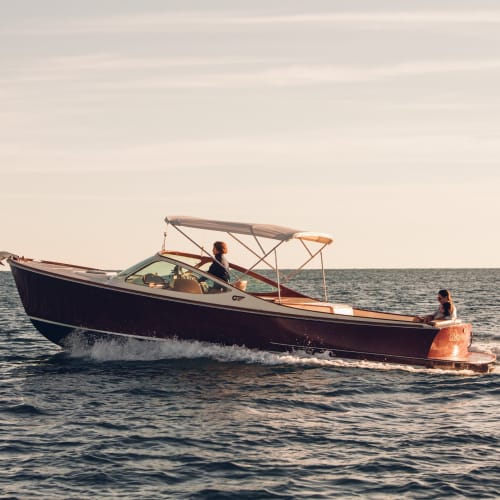 Ein Motorboot mit Dach, das bei Sonnenuntergang mit zwei Personen an Bord auf ruhigem Meer fährt, einer steuert und der andere sitzt hinten.
