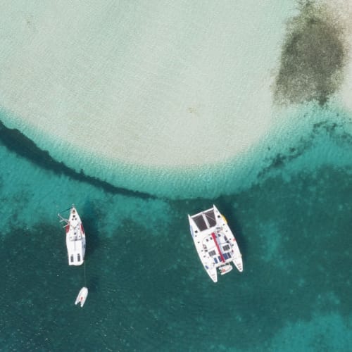 Zwei Segelboote und ein kleines Boot, die in klarem türkisfarbenem Wasser in der Nähe eines sandigen Ufers vor Anker liegen.